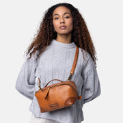 A woman with long curly brown hair models a small brown leather top-handle bag with a shoulder strap, worn crossbody over her chest. She is wearing a light gray knit sweater and white textured pants against a solid light gray background.