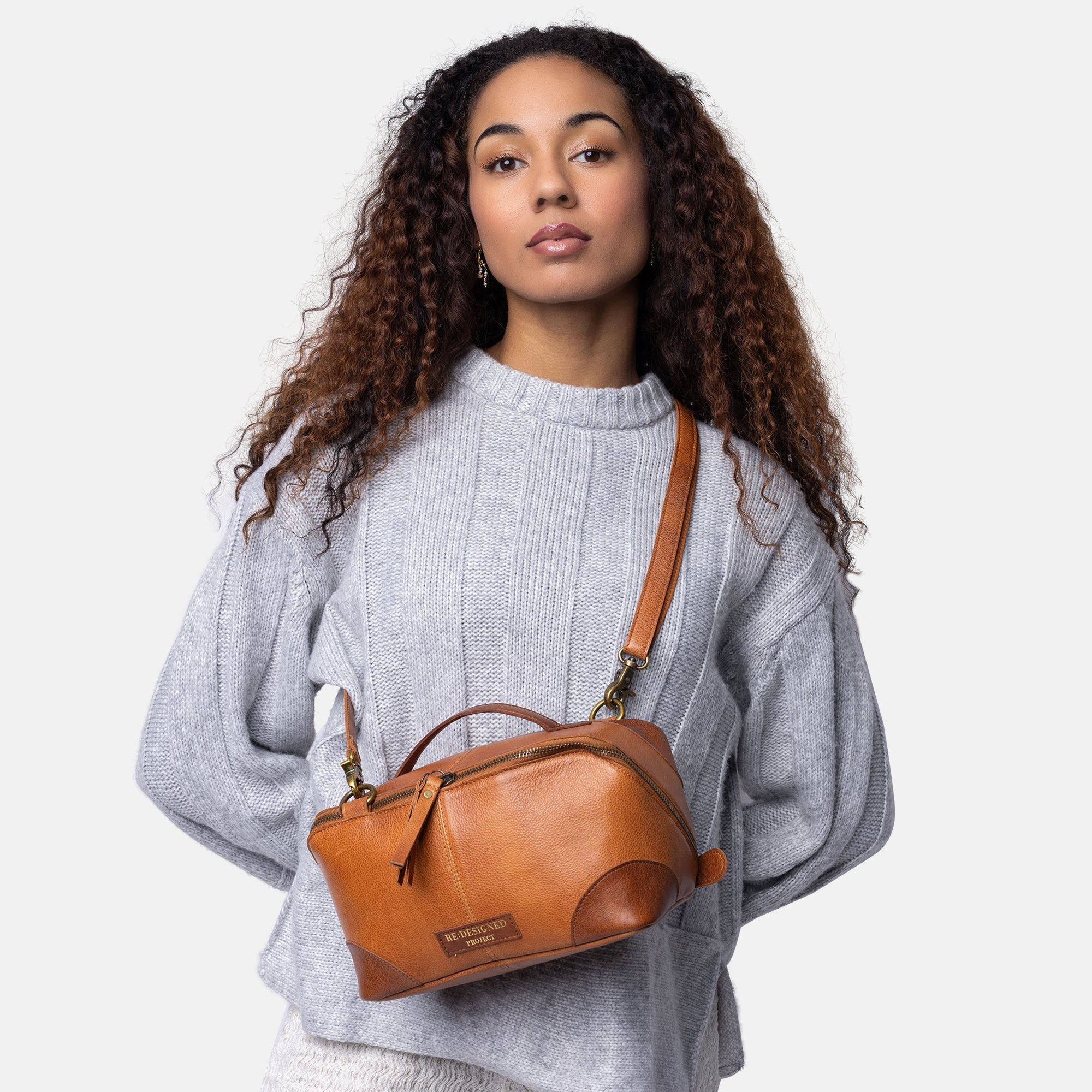 A woman with long curly brown hair models a small brown leather top-handle bag with a shoulder strap, worn crossbody over her chest. She is wearing a light gray knit sweater and white textured pants against a solid light gray background.