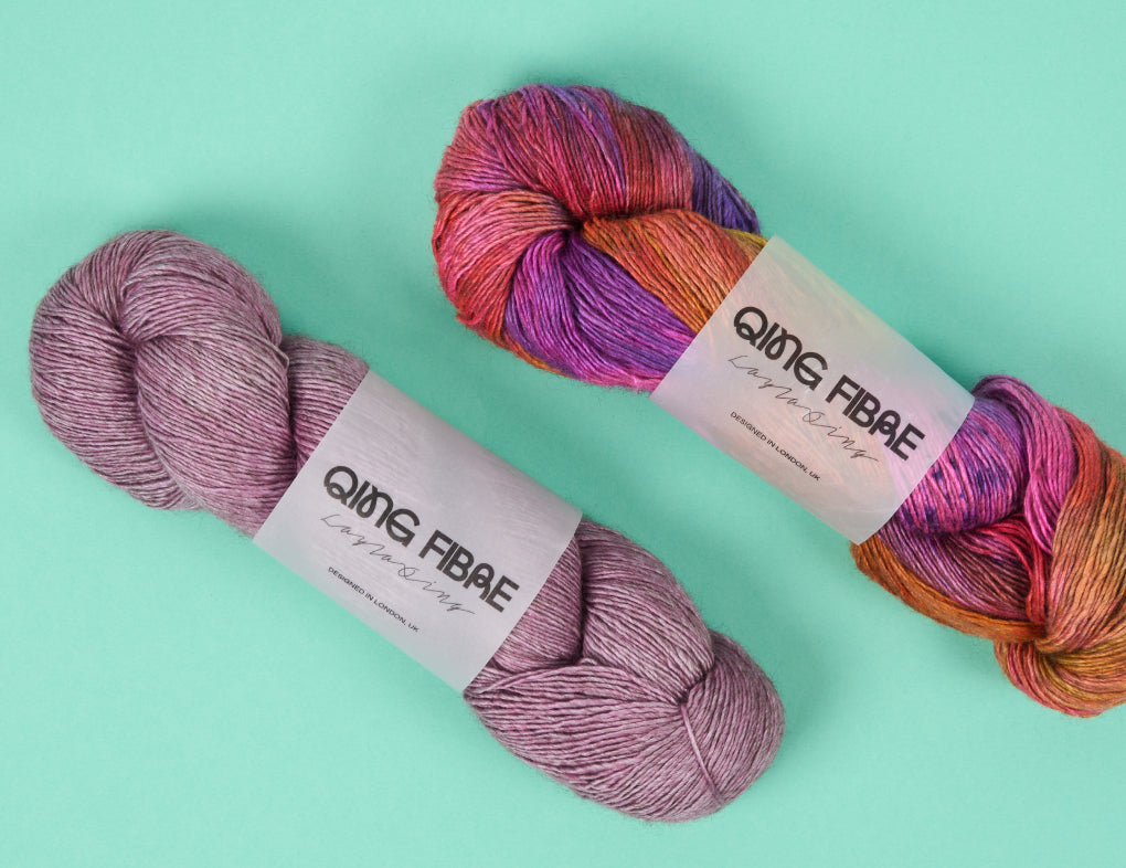 This image shows two skeins of Yak Singles from the brand Qing Fibre in warm pink, orange, and purple tones. They stand on a pale green background. 