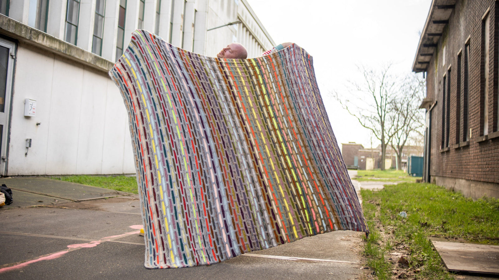 This image shows Stephen West wearing a beautifully knitted multicolour sample of the Painting Bricks Blanket from Westknits. 