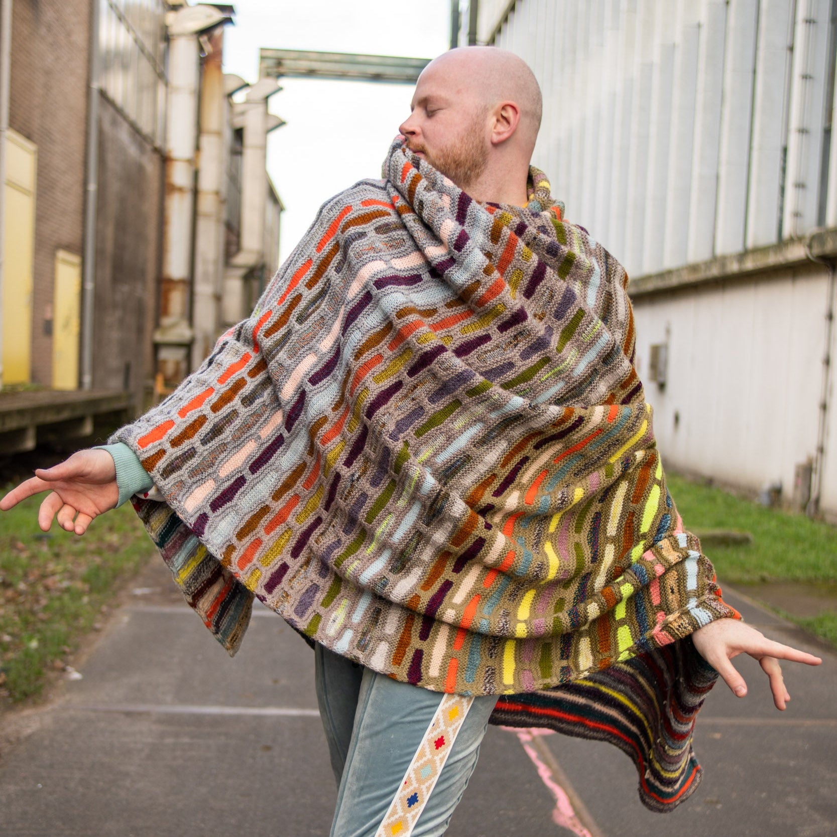 This image shows Stephen West wearing a beautifully knitted multicolour sample of the Painting Bricks Blanket from Westknits. 