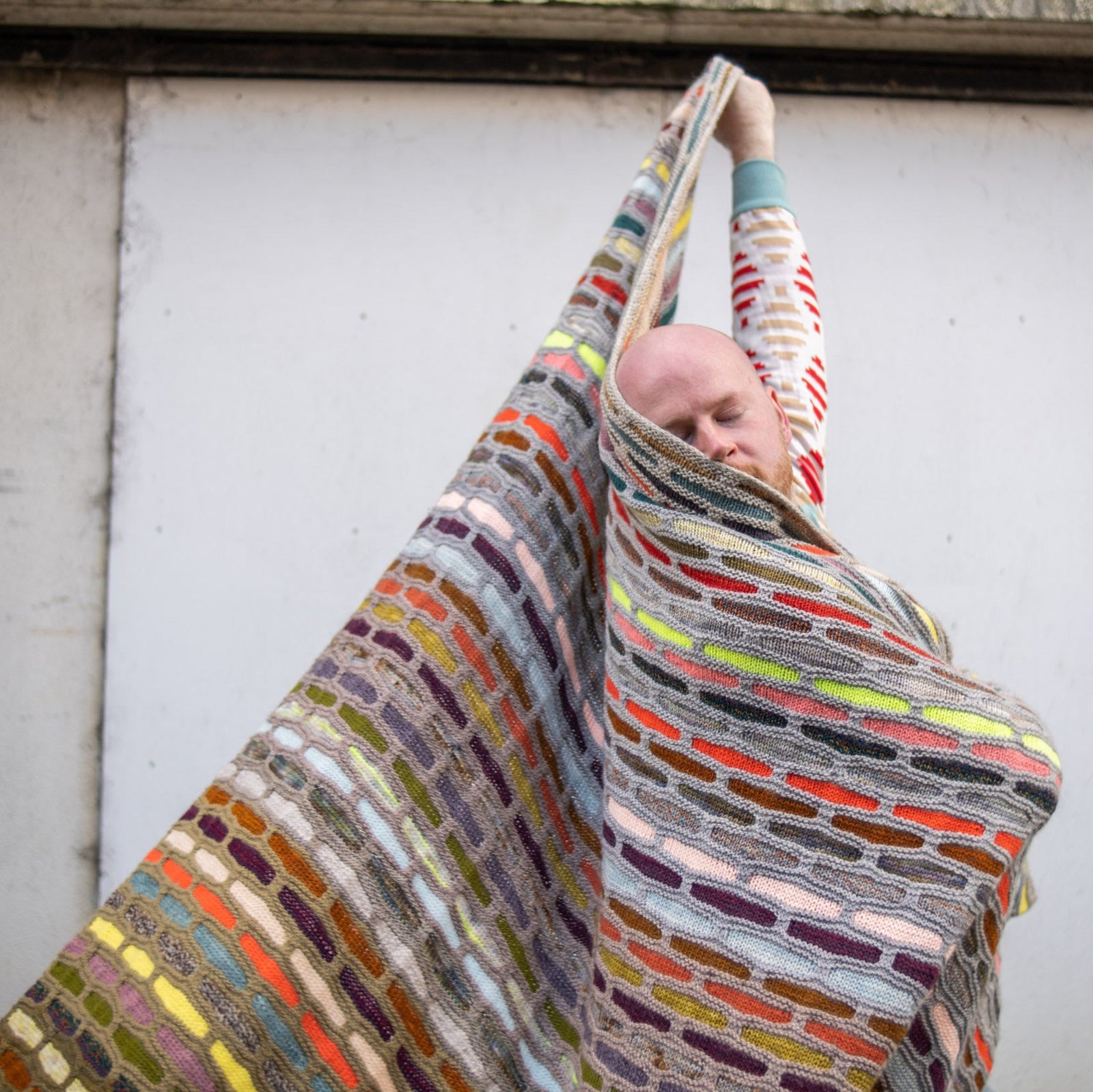 This image shows Stephen West wearing a beautifully knitted multicolour sample of the Painting Bricks Blanket from Westknits. 