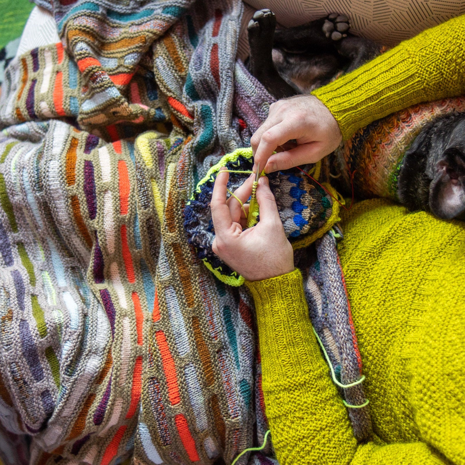 This image shows Stephen West knitting while wearing a beautifully knitted multicolour sample of the Painting Bricks Blanket from Westknits. 