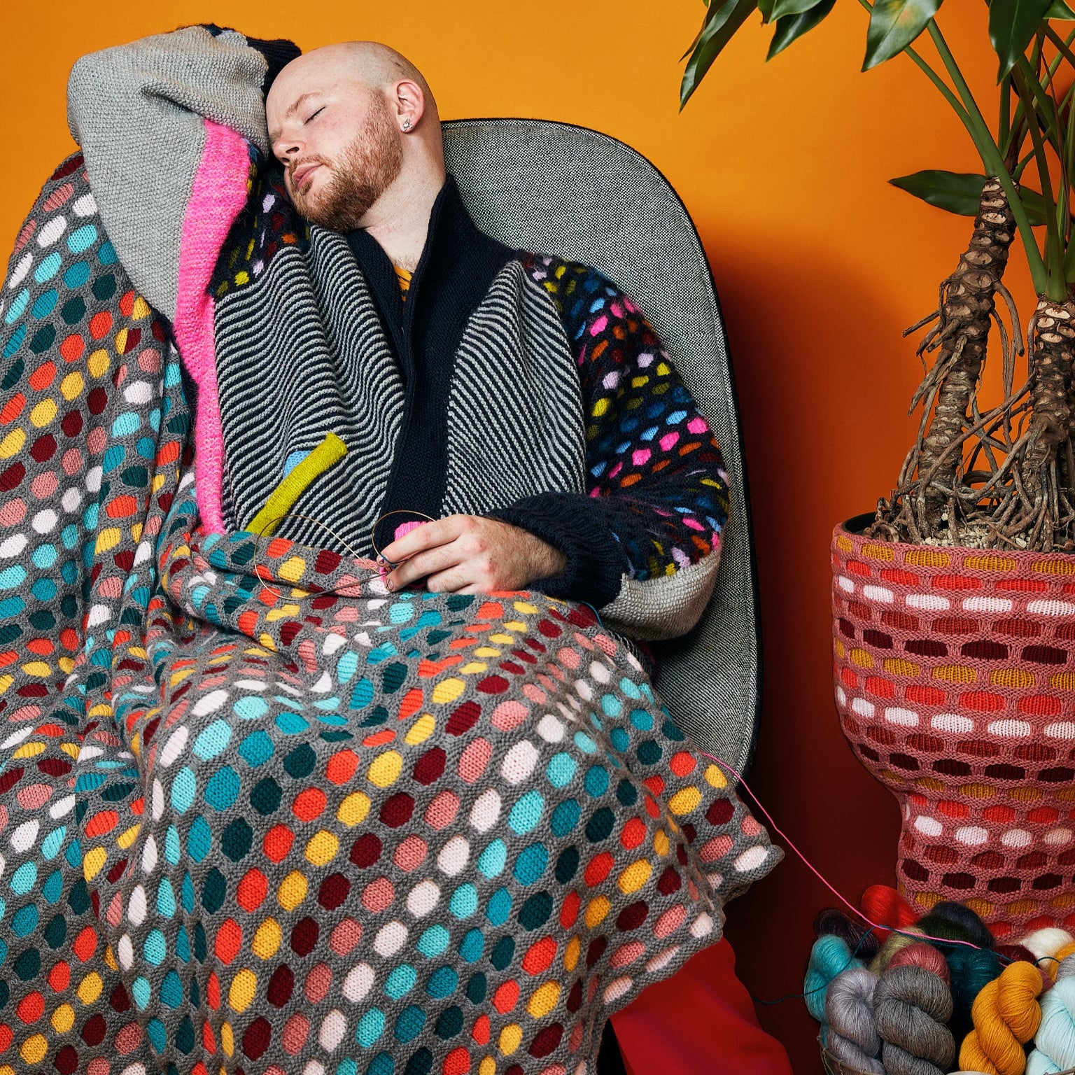 This image shows Stephen West wearing a knitted sample of the Painting Honeycombs Blanket by Westknits in various colours, including grey, yellow, orange, blue. 