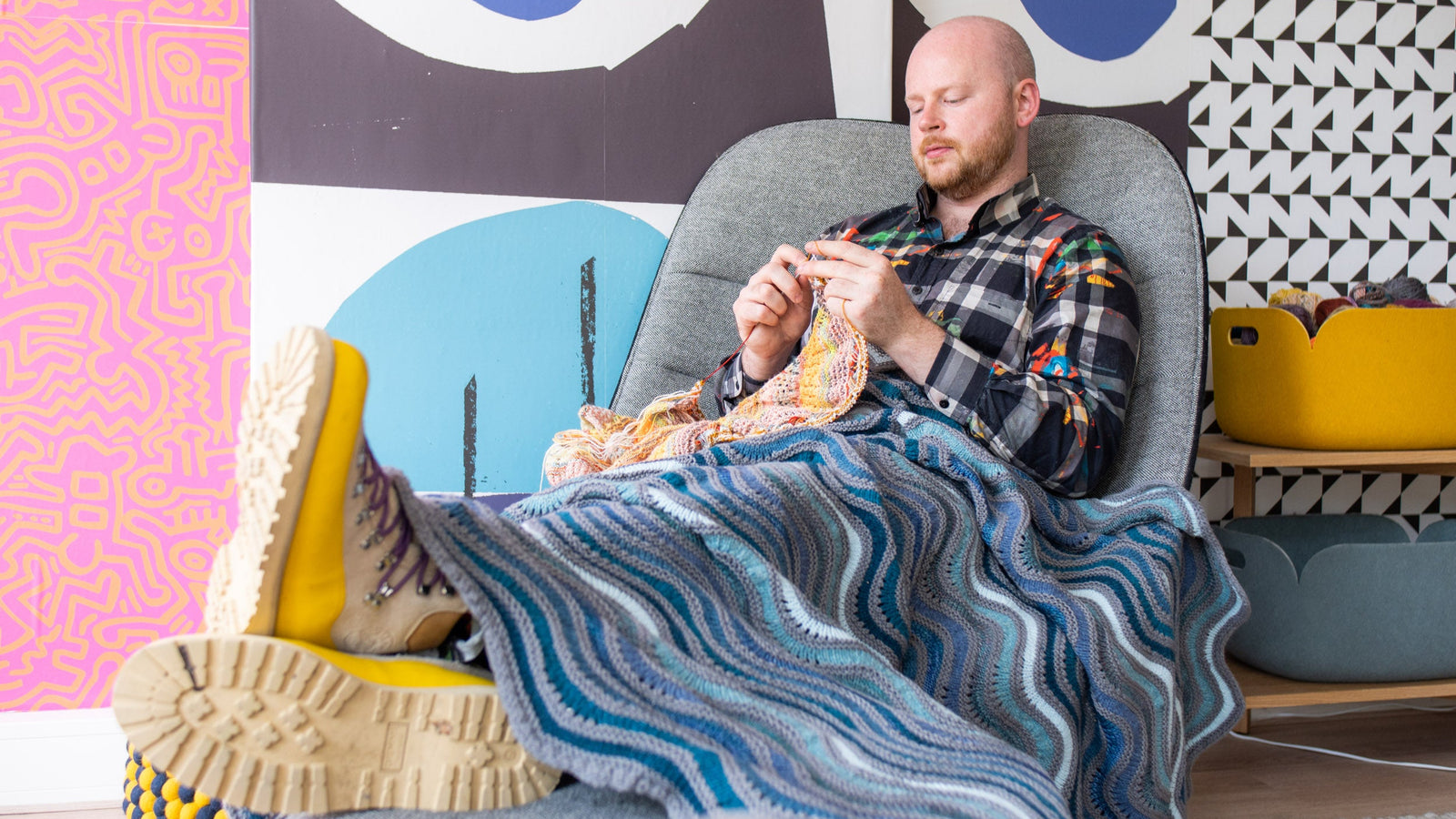This image shows Stephen West knitting while wearing a beautifully knitted sample of the Painting Waves Blanket in cool blue and grey tones. 