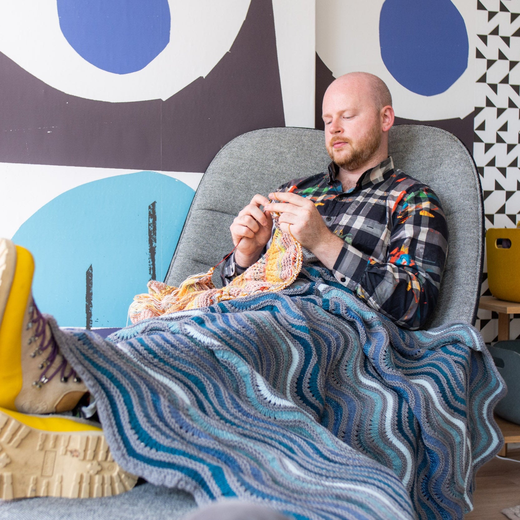 This image shows Stephen West knitting while wearing a beautifully knitted sample of the Painting Waves Blanket in cool blue and grey tones. 