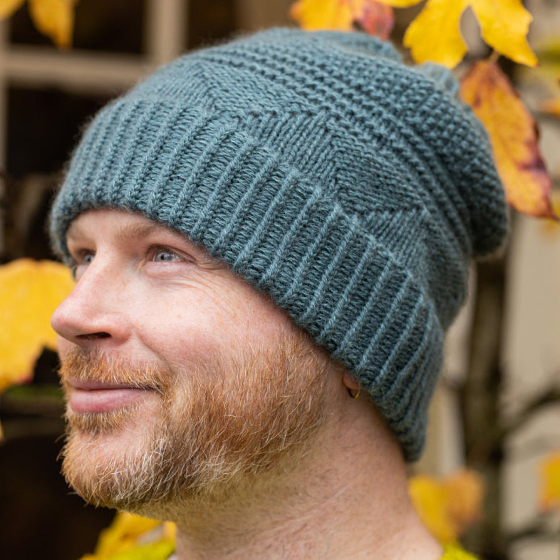 This image shows Stephen West wearing the Pierre Hat, which is knitted in a solid blue colour. The background shows yellow autumnal leaves. 
