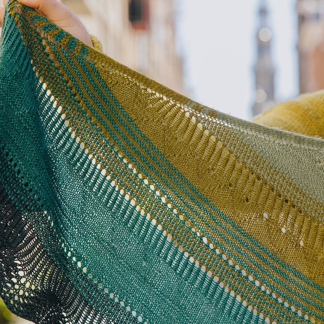 Stephen West holds up a Pierre Light knitted shawl with a gradient color story transitioning from deep green at one end, through deep teal and emerald green, to olive and mustard yellow at the other.