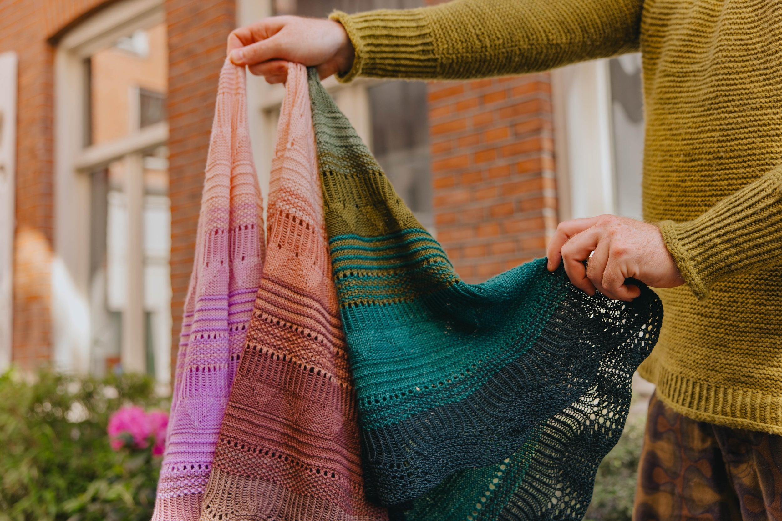 Stephen West holds up 3 Pierre Light shawls in lavender, dusty rose, and teal-green. 