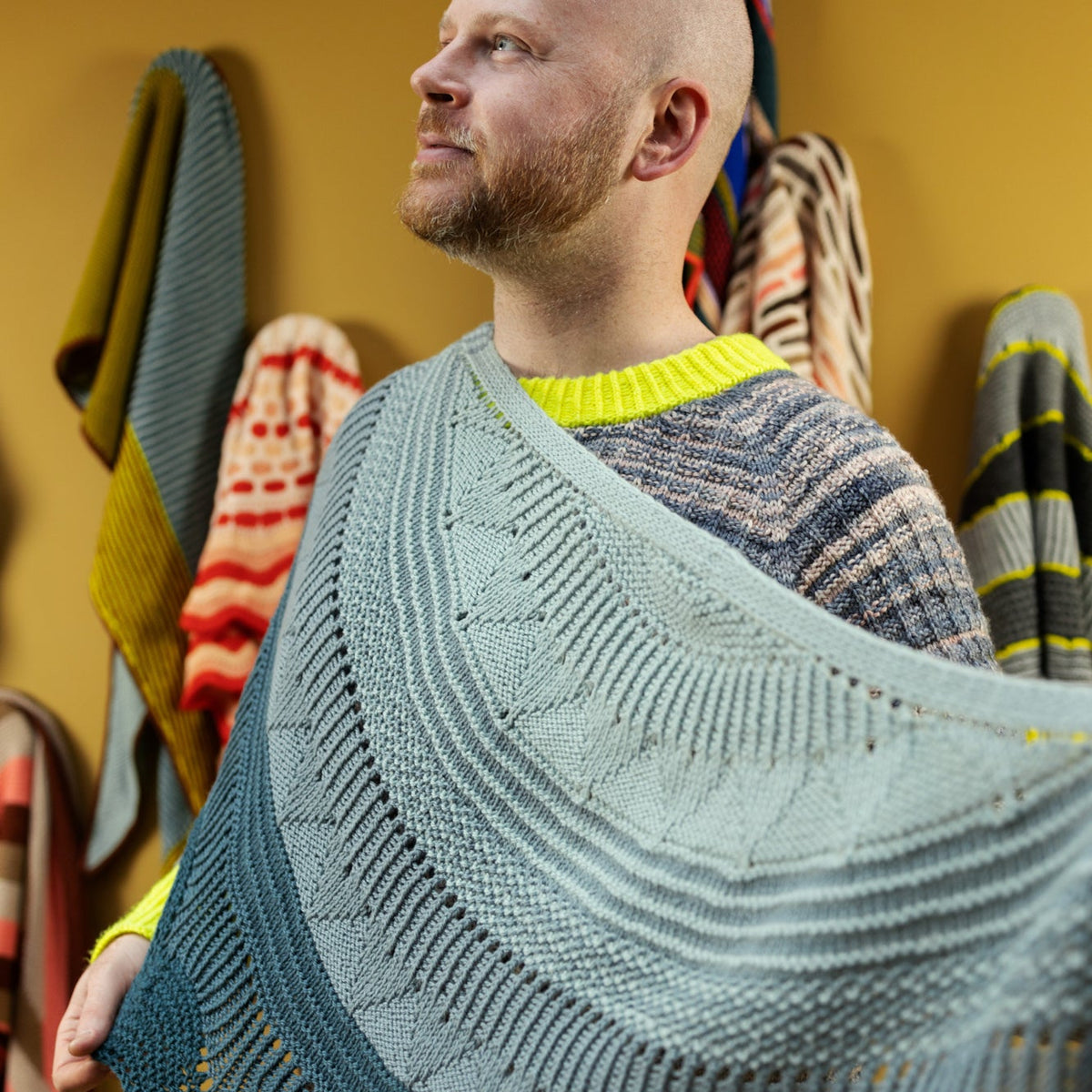 This image shows Stephen West wearing a knitted sample of the Pierre Shawl by Westknits in cool blue and grey tones. 