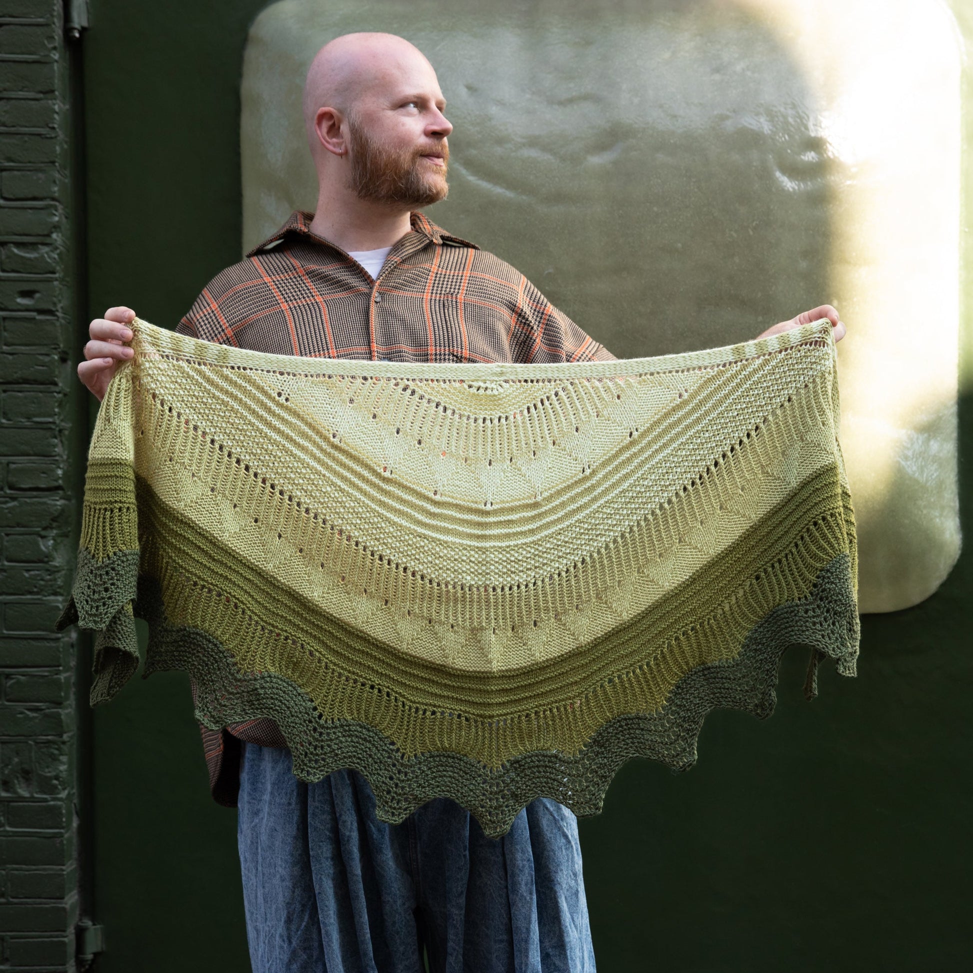 Stephen West holding a hand knitted sample of the Pierre shawl in green gradients in front of a green building.