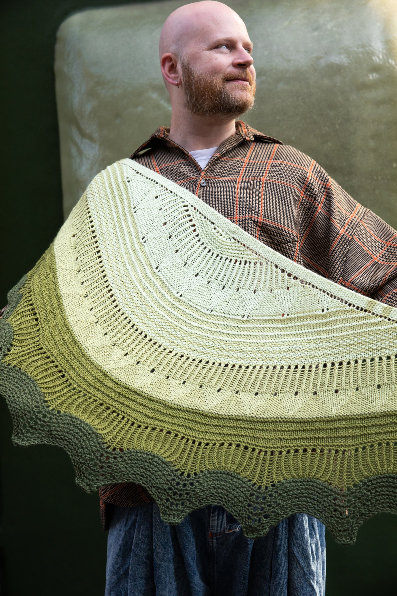 Stephen West wearing a hand knitted sample of the Pierre shawl in green gradients in front of a green building. 