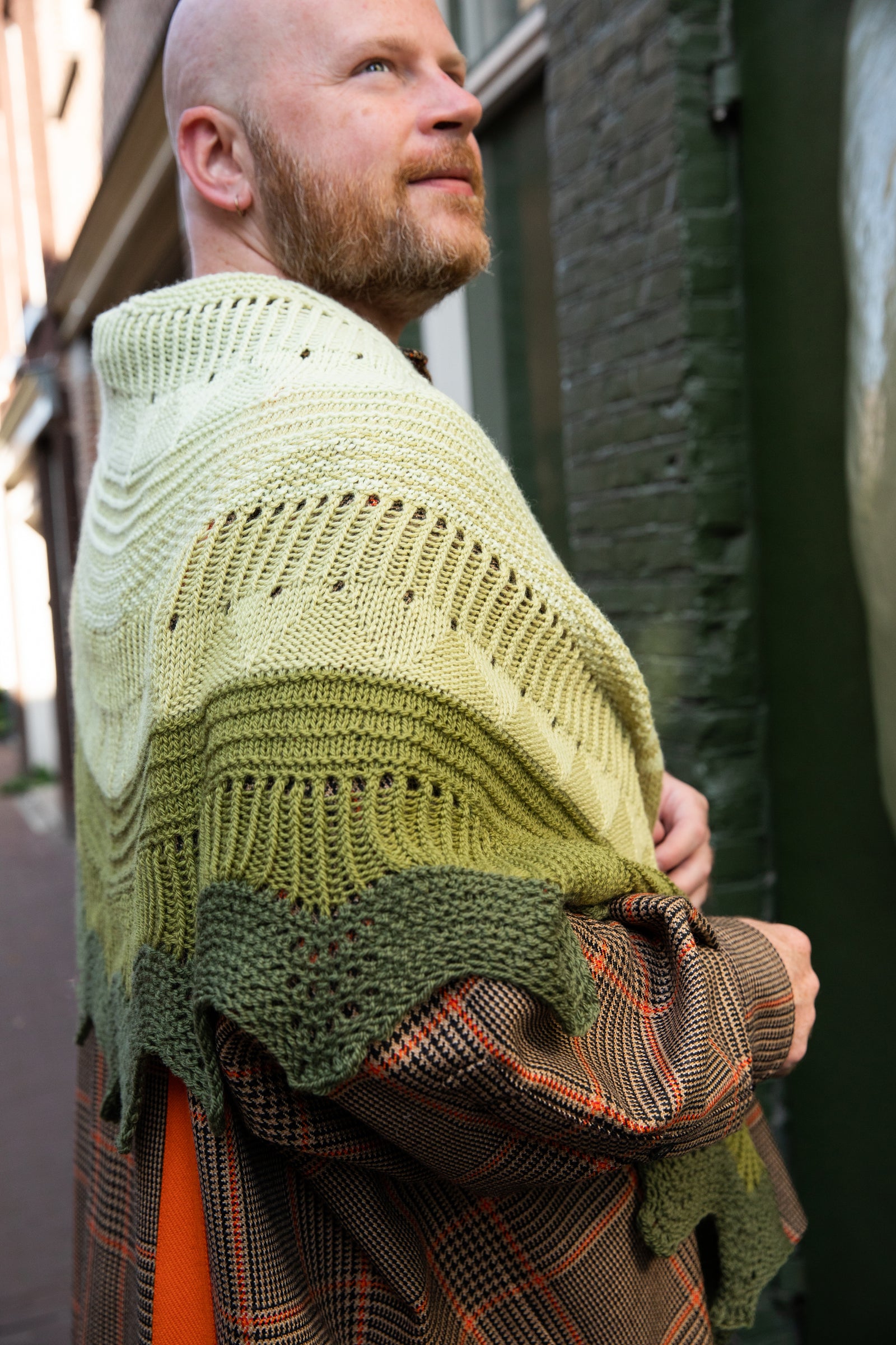 Stephen West wearing a hand knitted sample of the Pierre shawl in green gradients in front of a green building. 