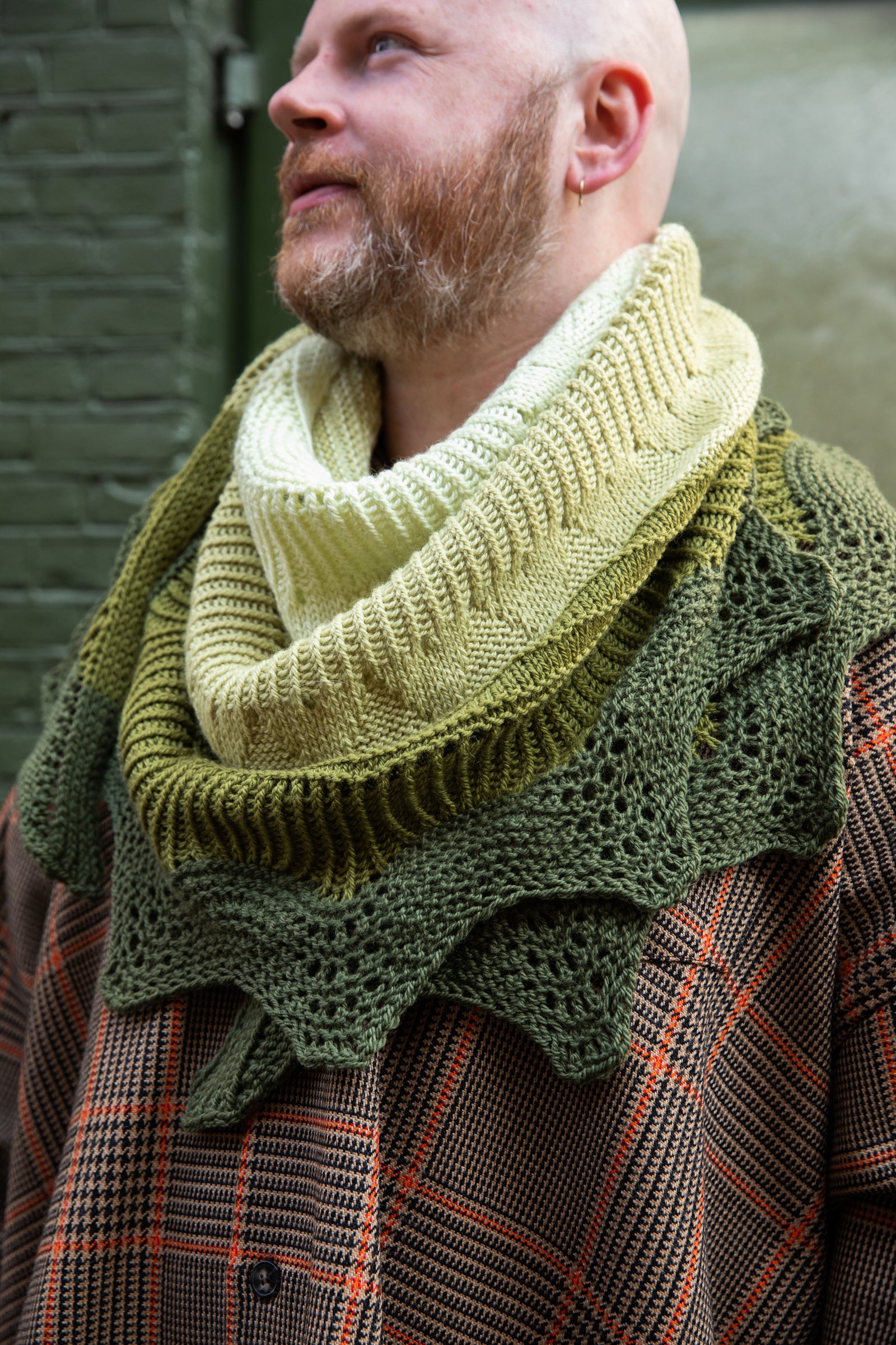 Stephen West wearing a hand knitted sample of the Pierre shawl in green gradients in front of a green building. 