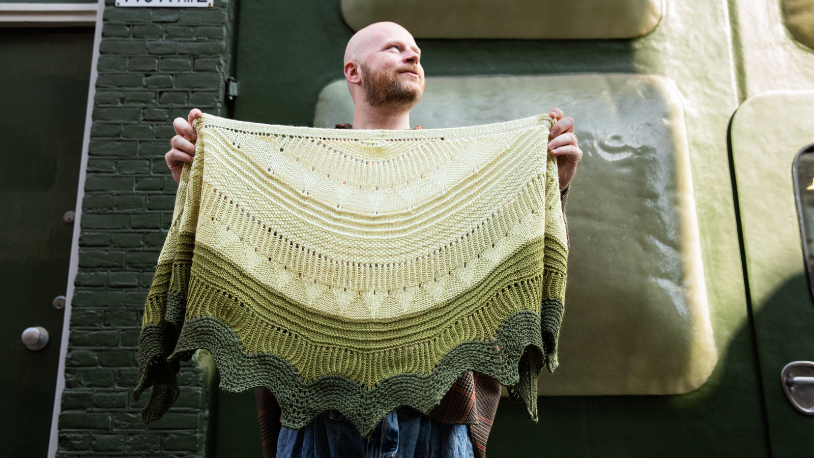 Stephen West holding a hand knitted sample of the Pierre shawl in green gradients in front of a green building. 