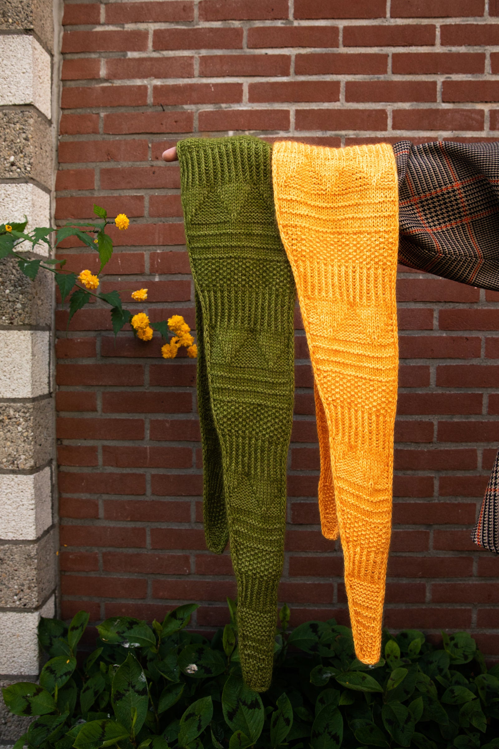 Stephen West holding two hand knitted samples of a skinny scarf in orange and green. 