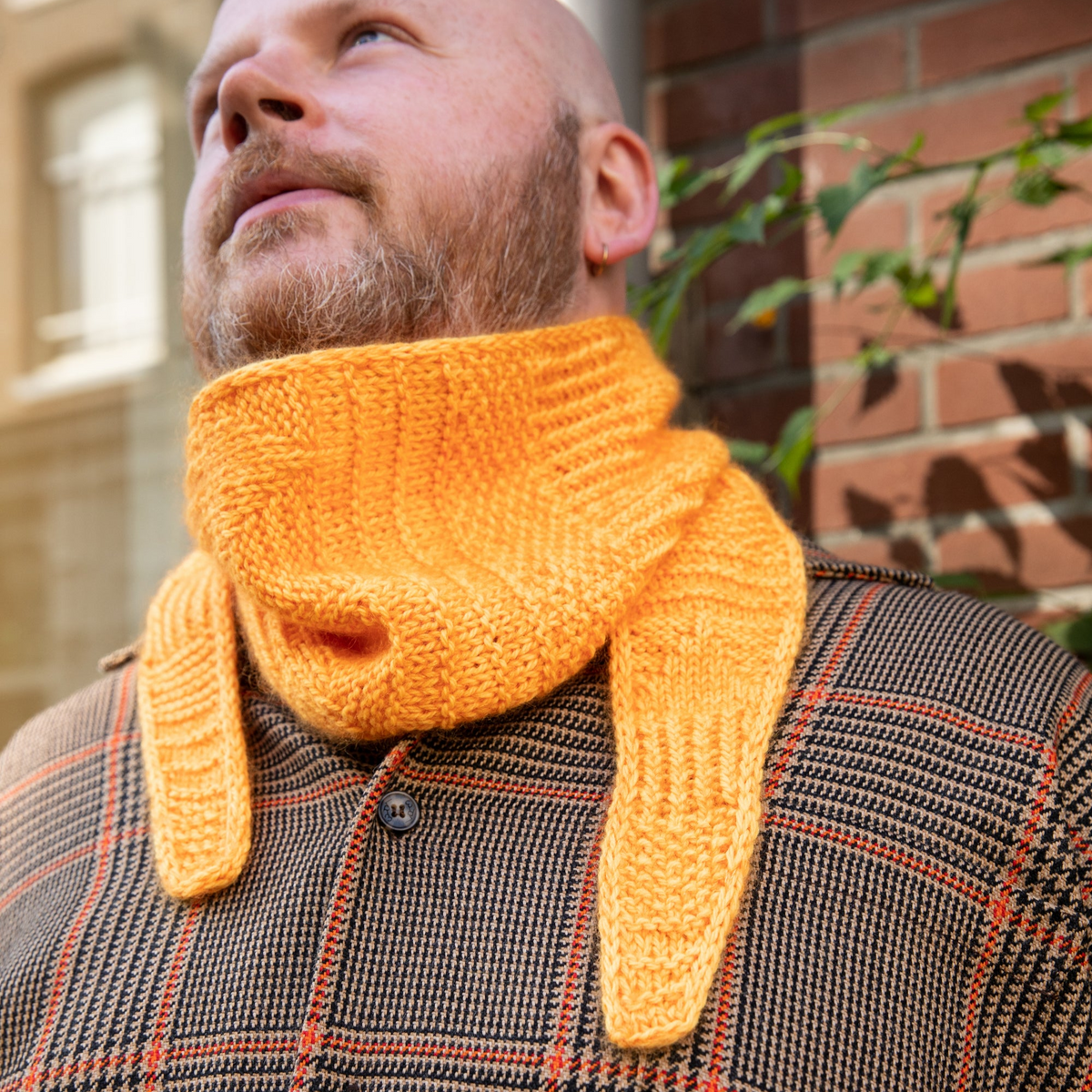 Stephen West wearing a hand knitted sample of an orange skinny scarf. 