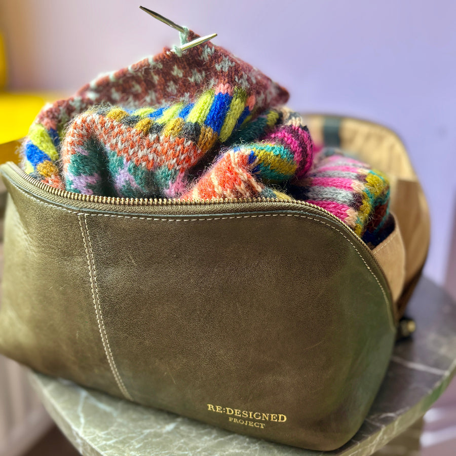 An olive green leather zippered pouch with gold 'REDESIGNED Project' branding, filled with a vibrant multicolor knit sweater, resting on a marble-topped stool.