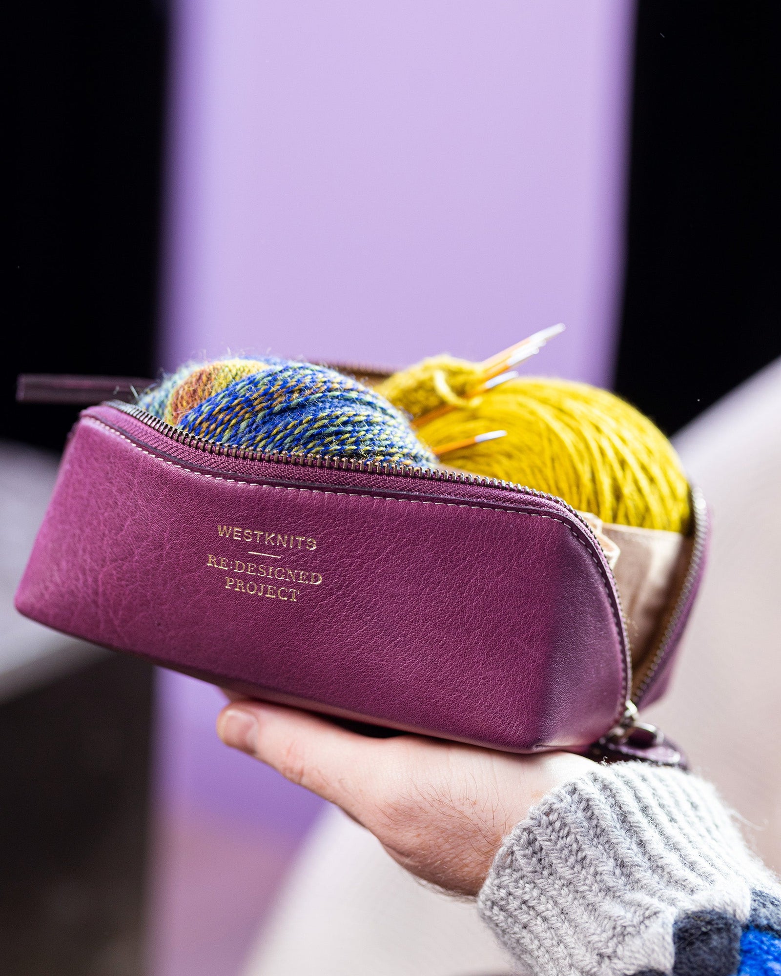 This image shows Project 36 - Aubergine, filled with yarn and needles. The bag is part of a collaboration between Re:Designed and Westknits. The bag stands on Stephen West's hand. 