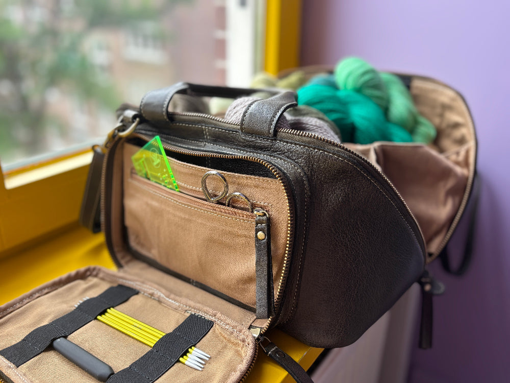 This image shows a dark green project bag from Re:Designed filled with West Wool yarn and notions, next to a window. 