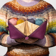 Stephen West, wearing a colorful striped knit sweater holds an open purple leather knitting bag filled with balls of yarn. The background is a clean, light gray studio backdrop.