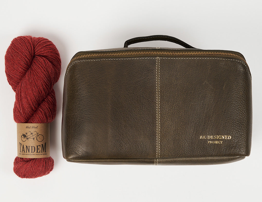 This image shows a dark green project bag from Re:Designed next to a skein of West Wool Tandem in red. 