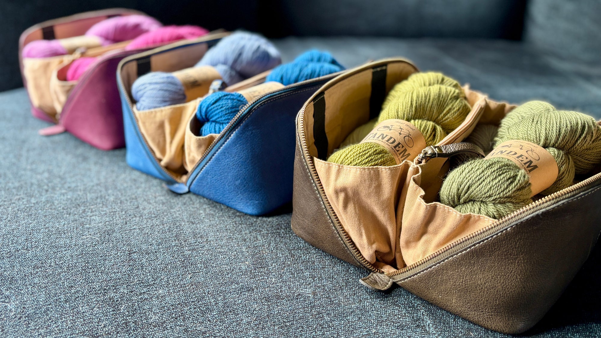 This image shows 3 project 9 in aubergine, denim, and dark green from Re:Designed, laid on a sofa and filled with matching yarn. 