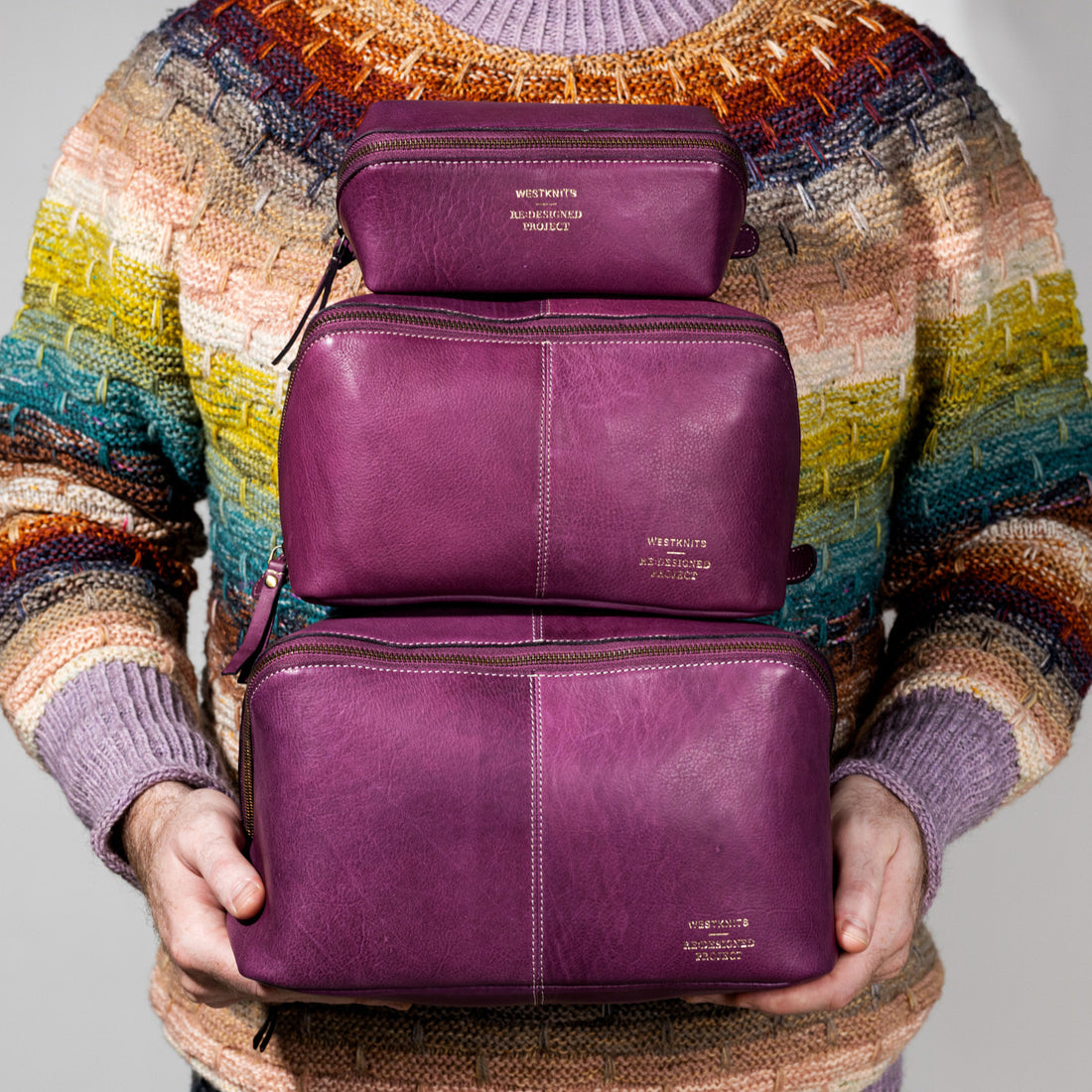 This image shows Stephen West holding three bags from the collaborations between Westknits and Re:Designed. The aubergine bags have the same style but in three different sizes. 
