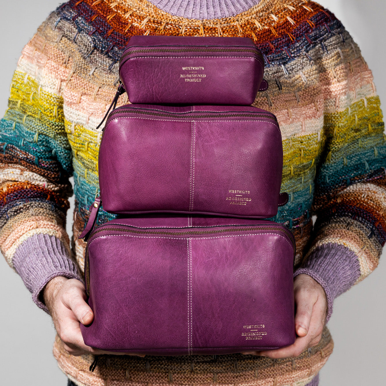 This image shows Stephen West holding three bags from the collaborations between Westknits and Re:Designed. The aubergine bags have the same style but in three different sizes. 