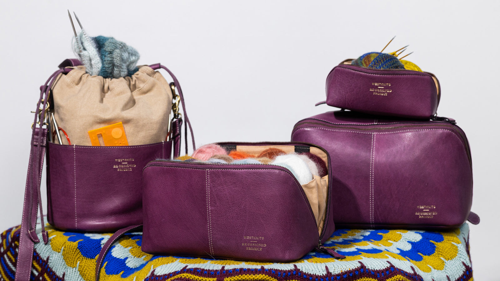 This image shows 4 aubergine project bags of the collaboration between Westknits and Re:Designed. The bags lay on a beautifully knitted blanket in ochre, blue, and aubergine colours and they are filled with yarn. 