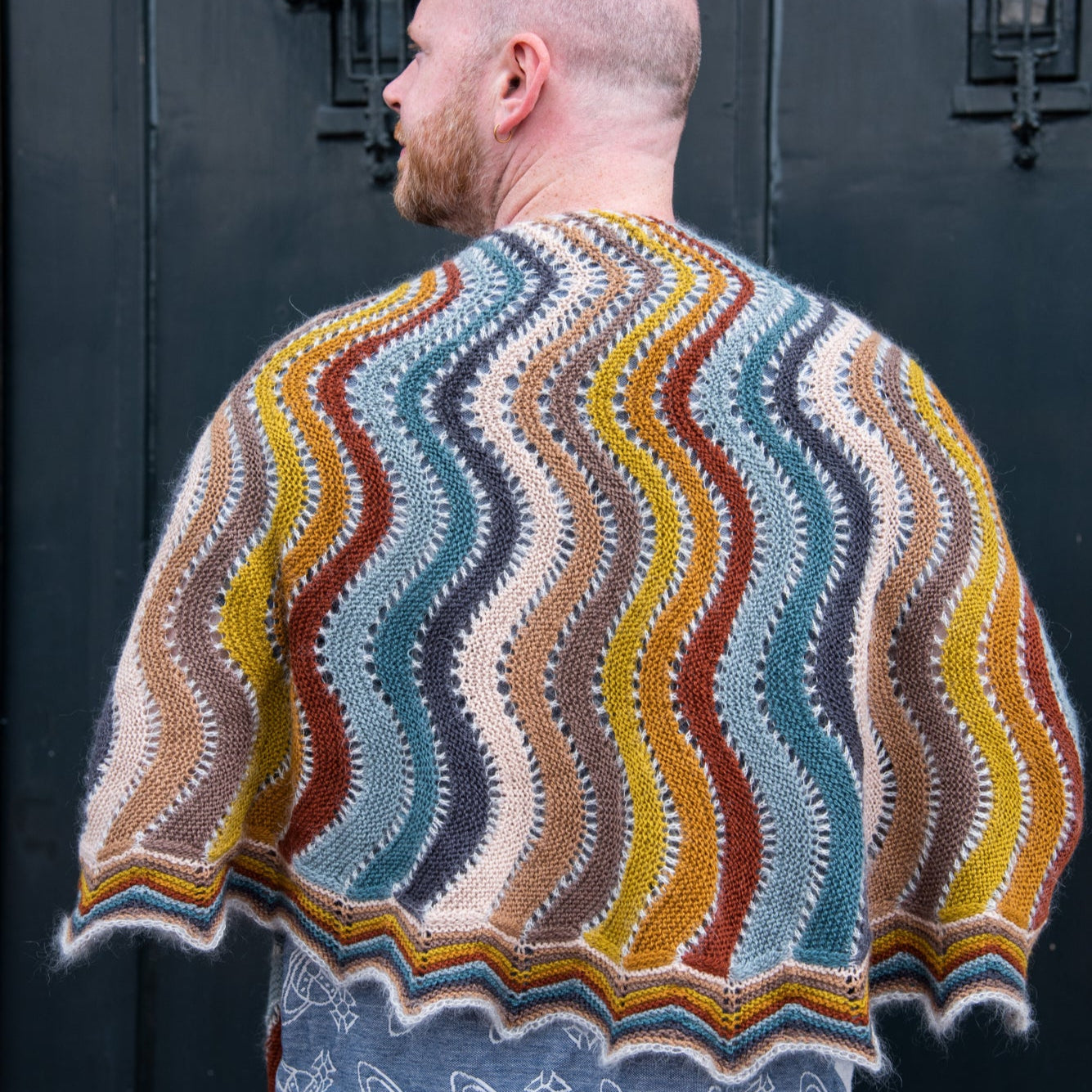 This image shows Stephen West wearing a beautifully knitted multicolour sample of the Replendence Shawl by Westknits. 