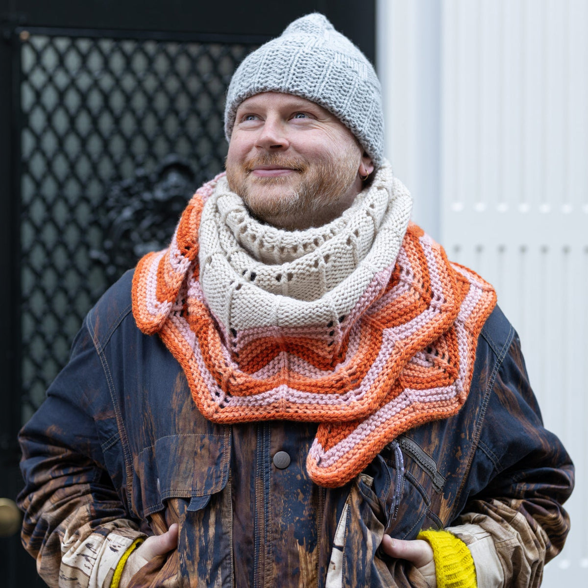 The image shows Stephen West wearing the Retro Chevron Shawl around his neck. The shawl is knitted with cream, orange, and pink tones. The background shows a house with a dark door. 