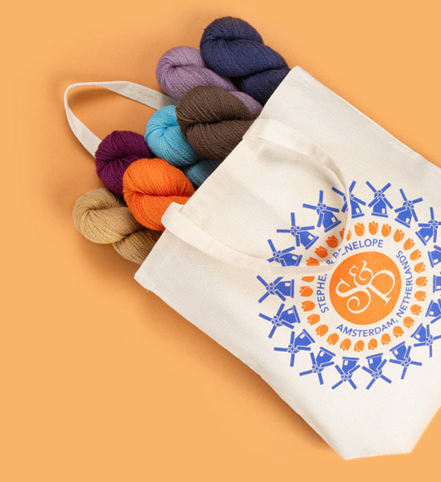 This image shows a canvas bag from Stephen & Penelope. Blue windmills and orange tulips are printed in a circle on the bag, as well as the S&P logo. 7 skeins of yarn in various colour, like orange, purple, and blue, come out from the bag. The items stand on a light orange background. 