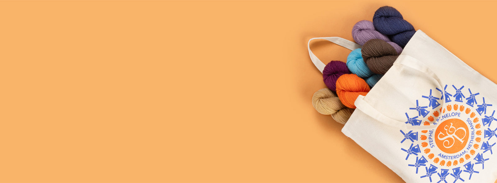 This image shows a canvas bag from Stephen & Penelope. Blue windmills and orange tulips are printed in a circle on the bag, as well as the S&P logo. 7 skeins of yarn in various colour, like orange, purple, and blue, come out from the bag. The items stand on a light orange background. 