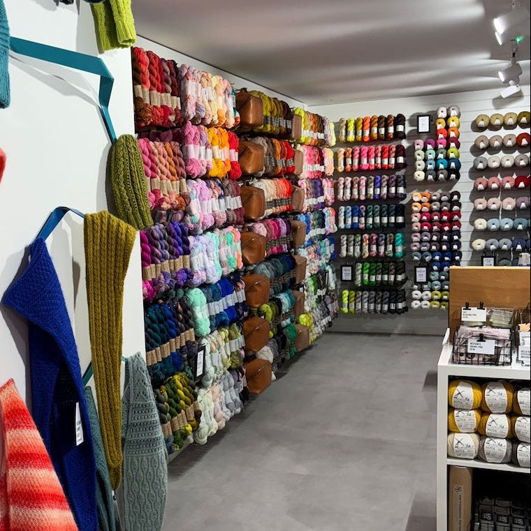 A display of colorful knitted hats and scarves hanging on a white wall in a yarn and knit shop, with shelves of yarn visible in the background.