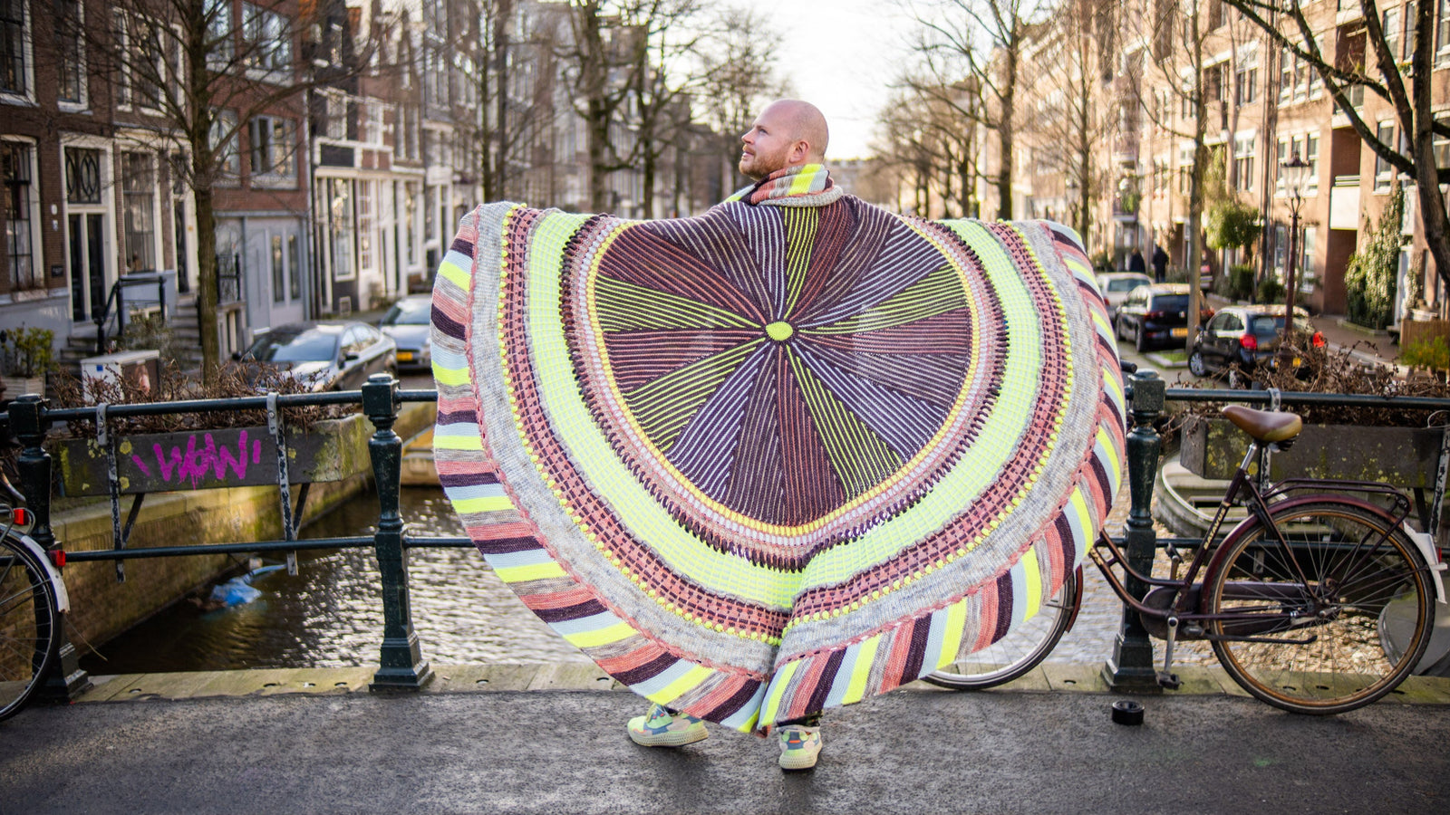 This image shows Stephen West wearing a beautifully knitted sample of the Shawlography Blanket by Westknits in purple, neon yellow, and coral tones.