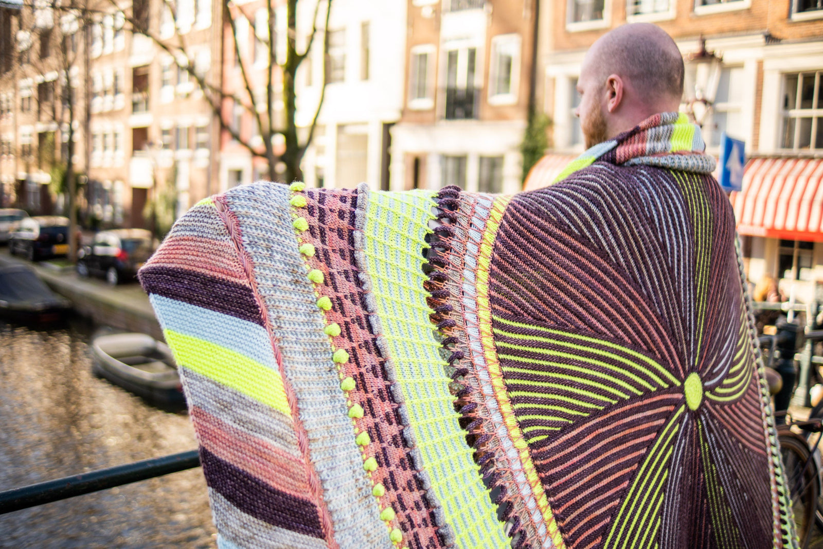 This image shows Stephen West wearing a beautifully knitted sample of the Shawlography Blanket by Westknits in purple, neon yellow, and coral tones.