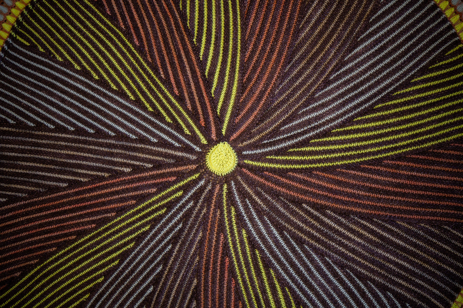 This image shows a detail of a beautifully knitted sample of the Shawlography Blanket by Westknits in purple, neon yellow, and coral tones.