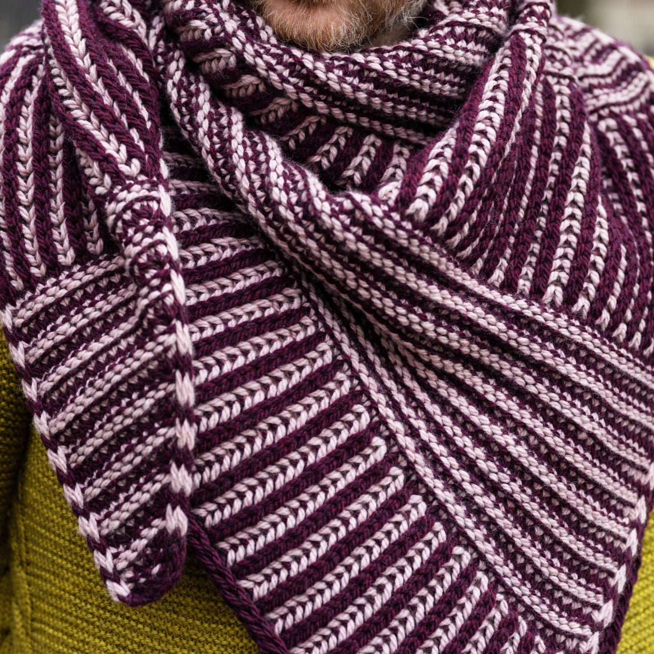  The image shows Stephen West wearing a sample of the Slip 'n Slide shawl by Westknits knit in purple and pink tones. The background shows a street of Amsterdam. 