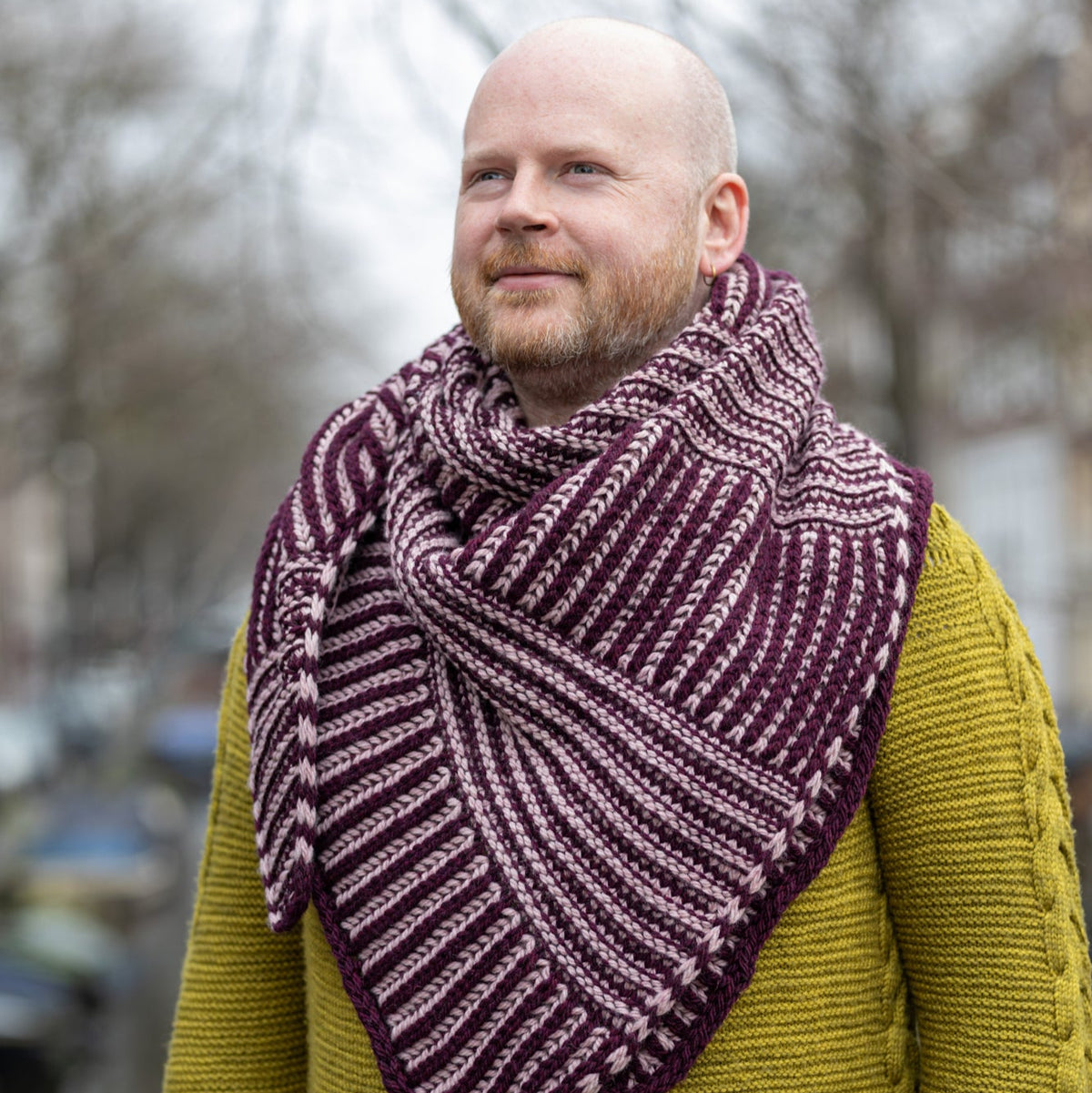 This image shows Stephen West wearing around his neck a beautifully knitted sample of the Slip 'n Slide Triangle in contrasting purple and pink tones. The background shows a canal in Amsterdam. 