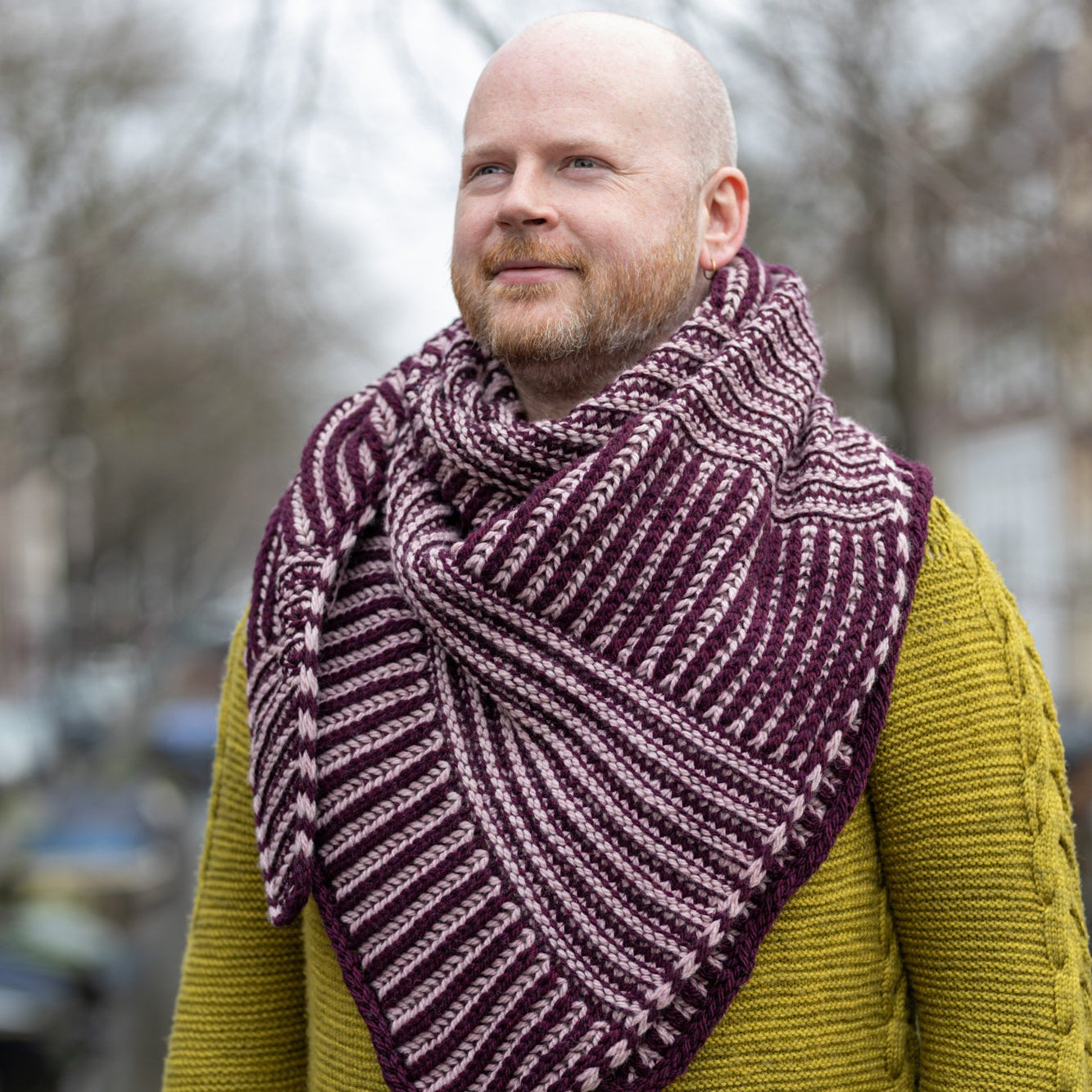  The image shows Stephen West wearing a sample of the Slip 'n Slide shawl by Westknits knit in purple and pink tones. The background shows a street of Amsterdam. 