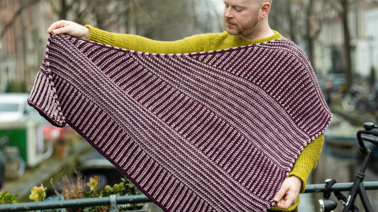  The image shows Stephen West holding a sample of the Slip 'n Slide shawl by Westknits knit in purple and pink tones. The background shows a street of Amsterdam. 