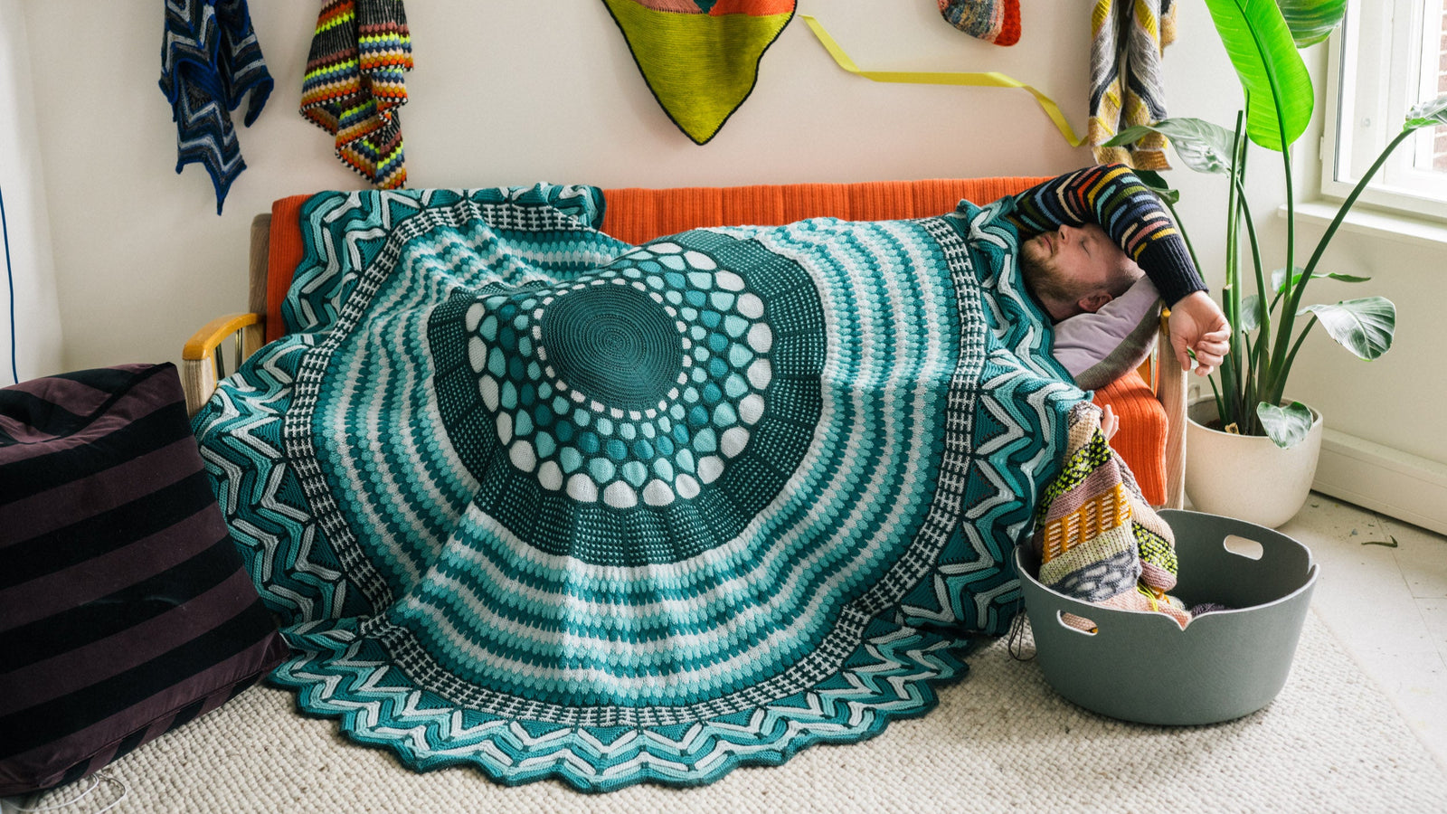 This image shows Stephen West sleeping under a beautifully knitted sample of the Slipstravaganza Blanket in cool blue and turquoise tones. 