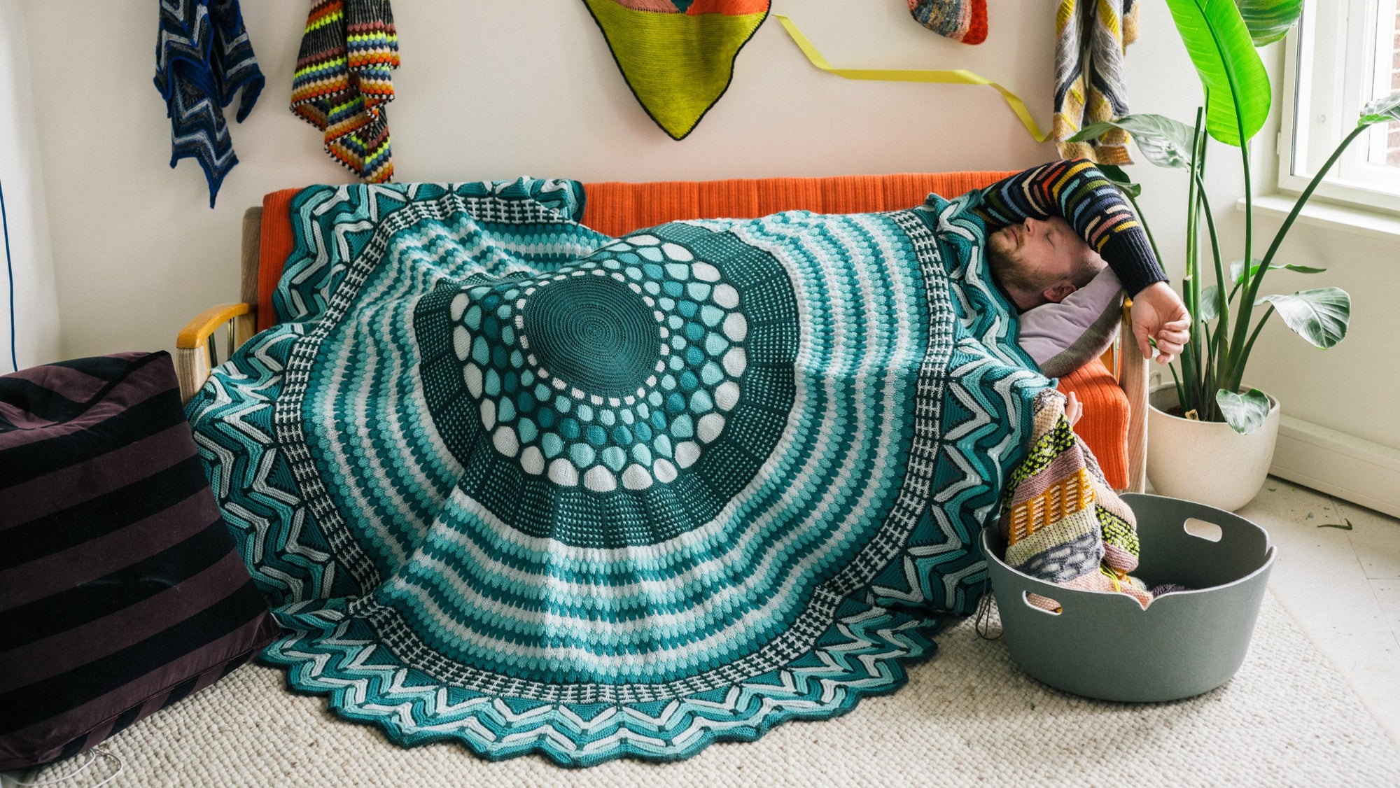 This image shows Stephen West sleeping under a beautifully knitted sample of the Slipstravaganza Blanket in cool blue and turquoise tones. 