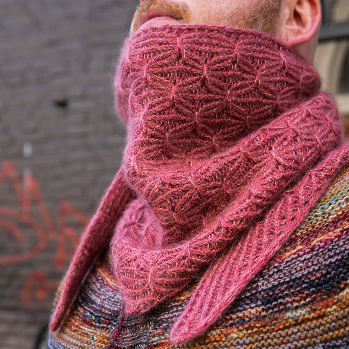 Stephen West wearing a hand knitted sample of a small shawl in red-pink tones. 