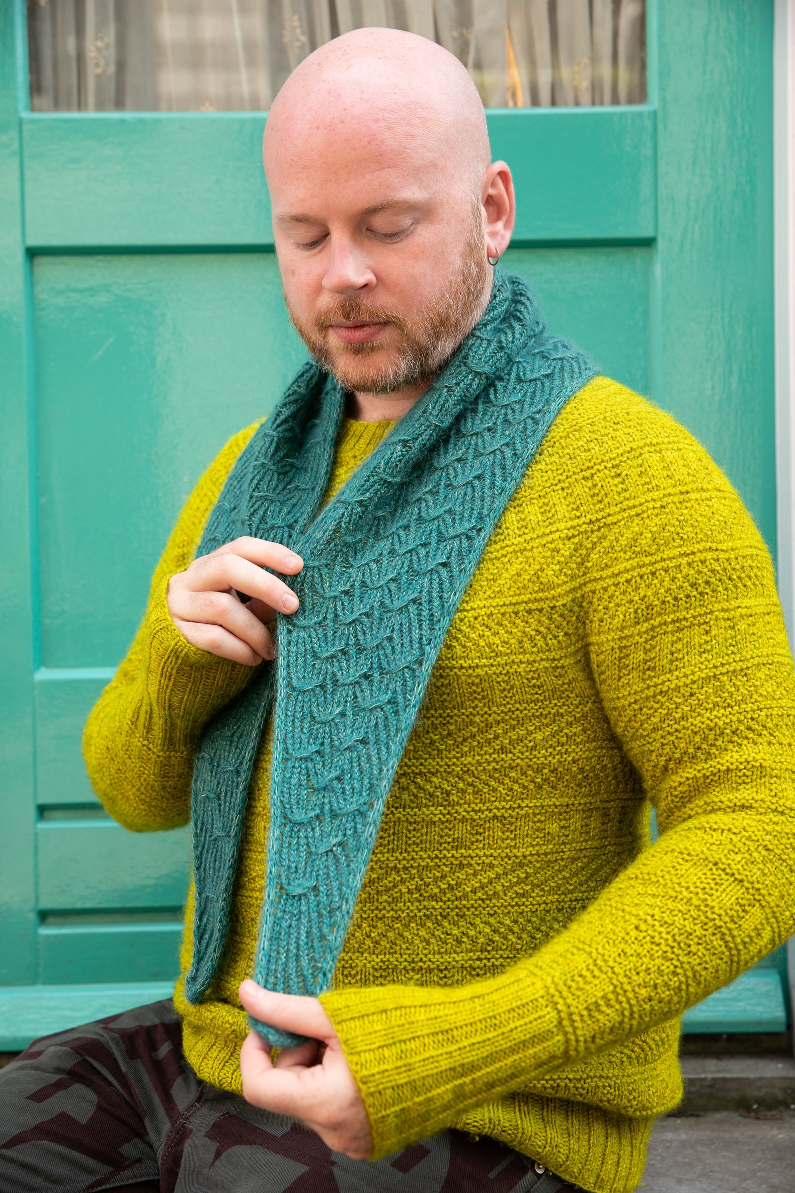 Stephen West wearing a hand knitted sample of a small shawl in blue-green tones. 