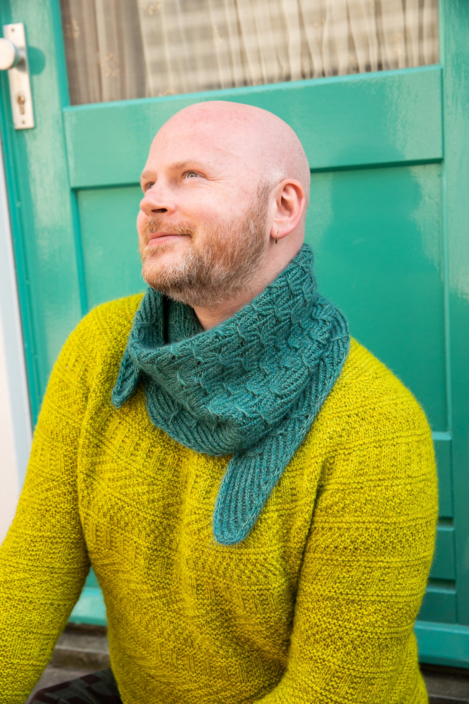 Stephen West wearing a hand knitted sample of a small shawl in blue-green tones. 
