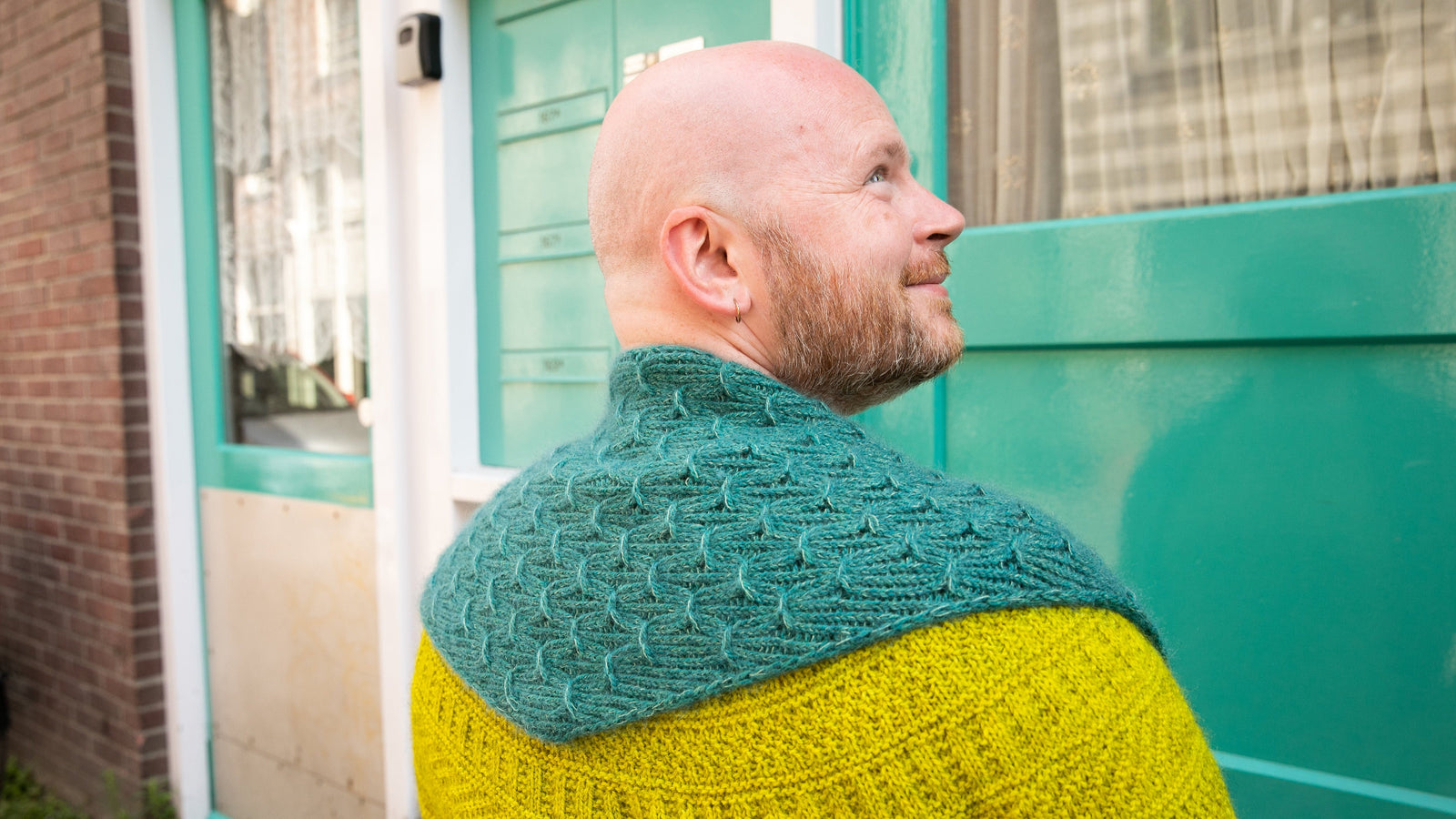 Stephen West wearing a hand knitted sample of a small shawl in blue-green tones. 