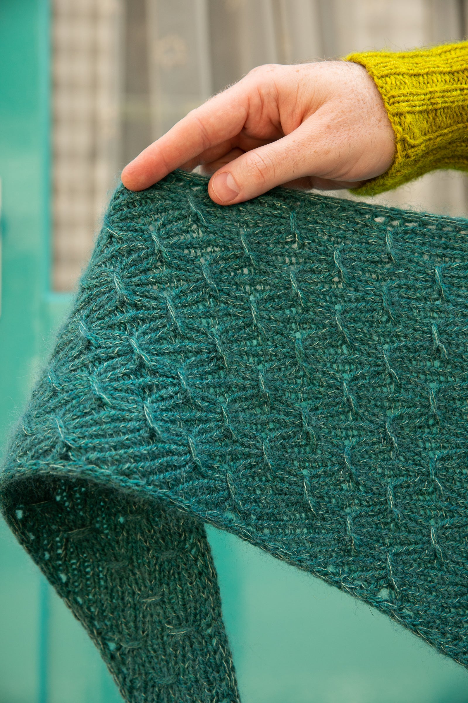 Stephen West holding a hand knitted sample of a small shawl in blue-green tones. 