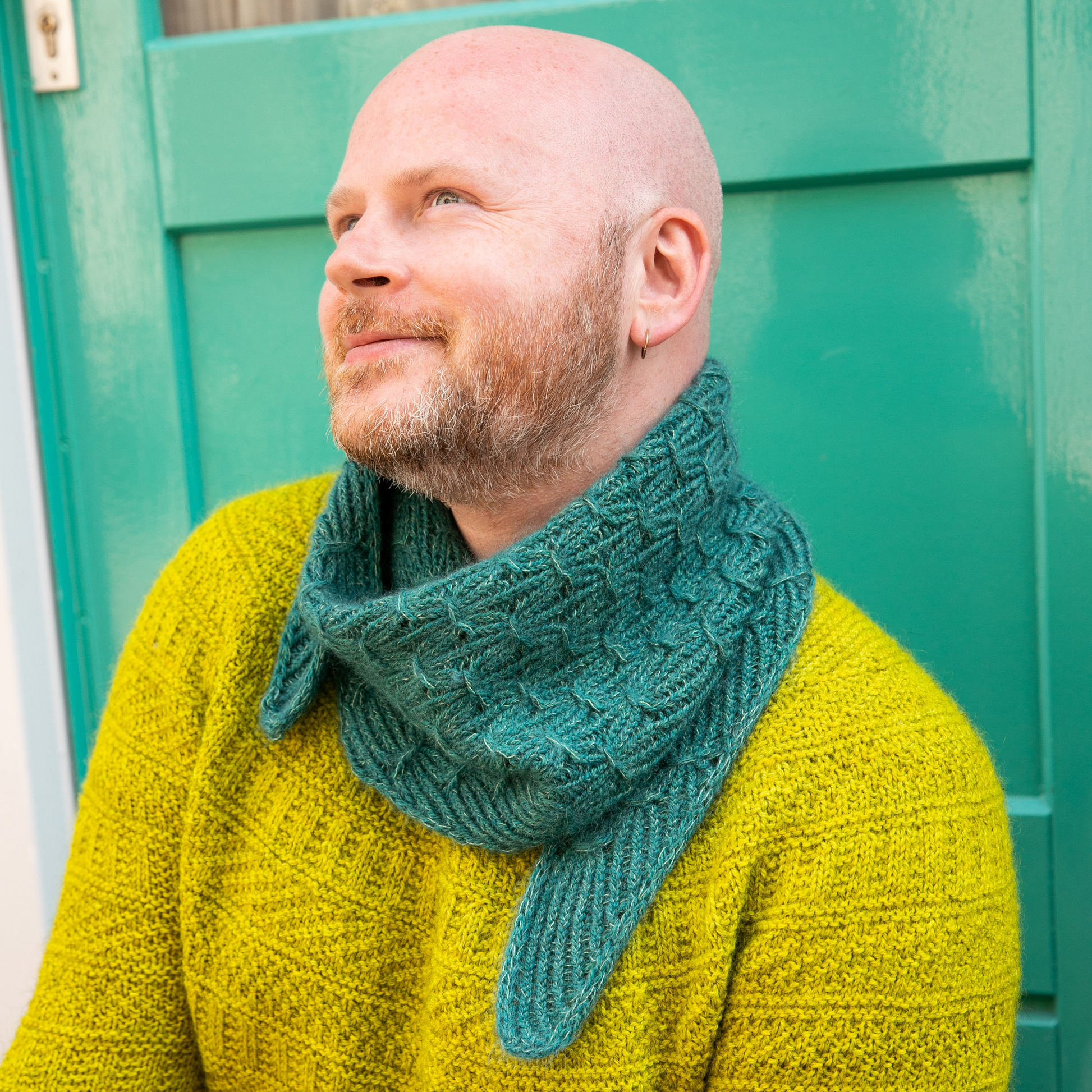 Stephen West wearing a hand knitted sample of a small shawl in blue-green tones.